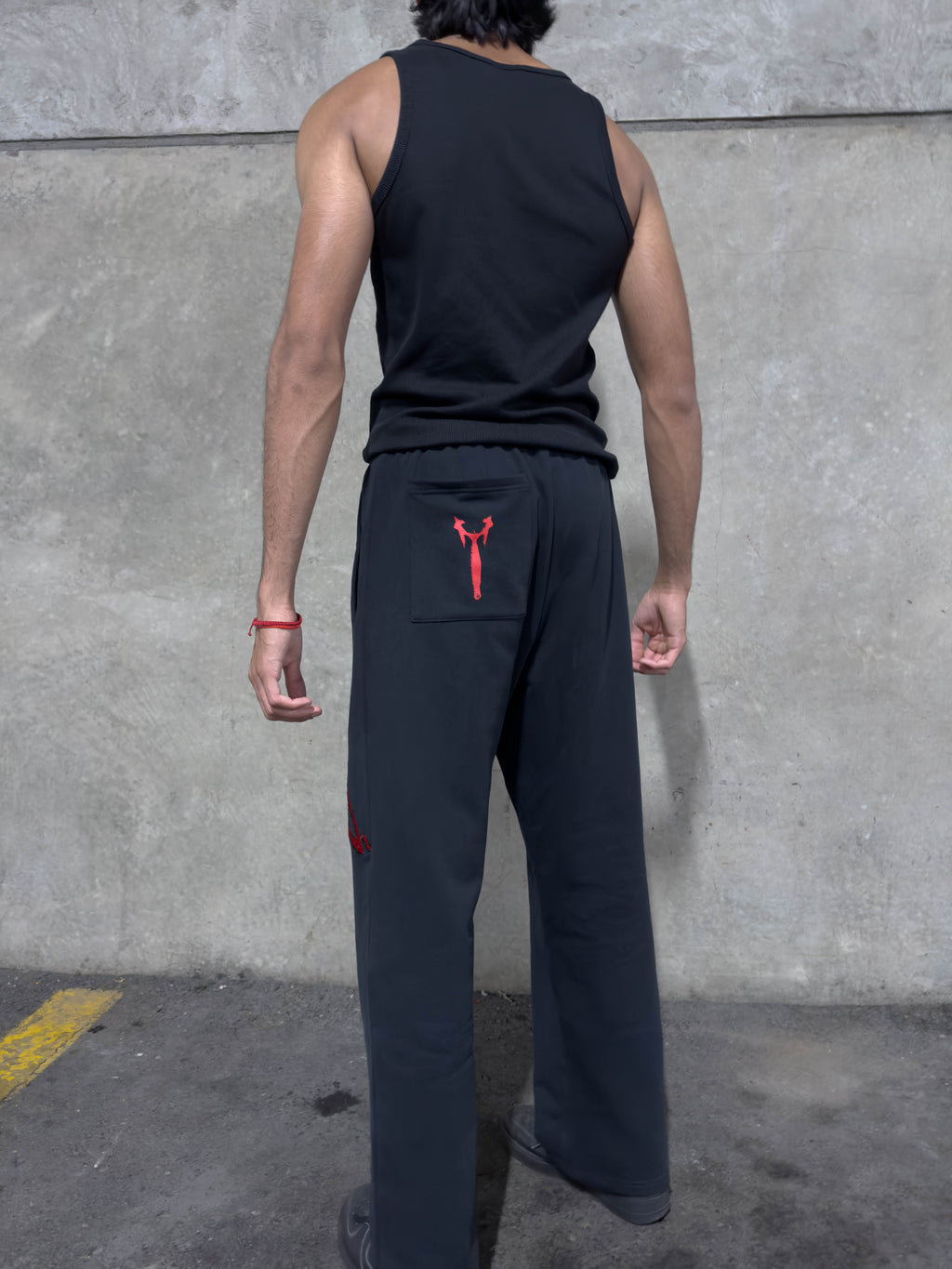 A man viewed from behind wearing black wide-leg sweatpants with a red drawstring and a red embroidered logo on the back pocket, paired with a black tank top against a concrete wall.