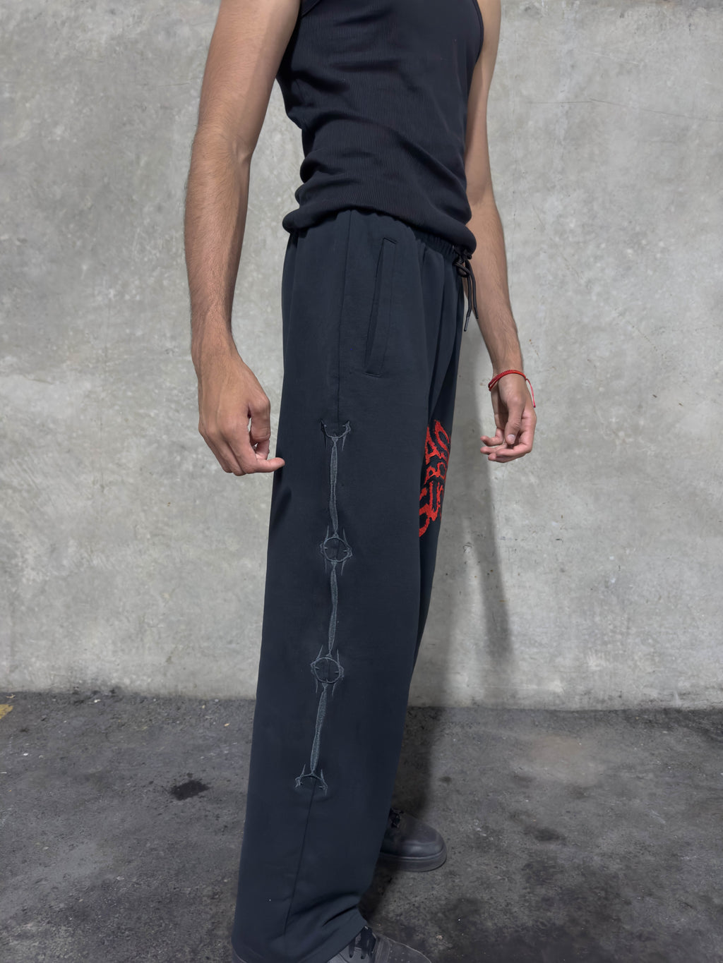 A person wearing loose black drawstring sweatpants with a vertical zipper detail and a red spider graphic on the thigh, paired with a black sleeveless top, standing against a textured gray concrete wall.