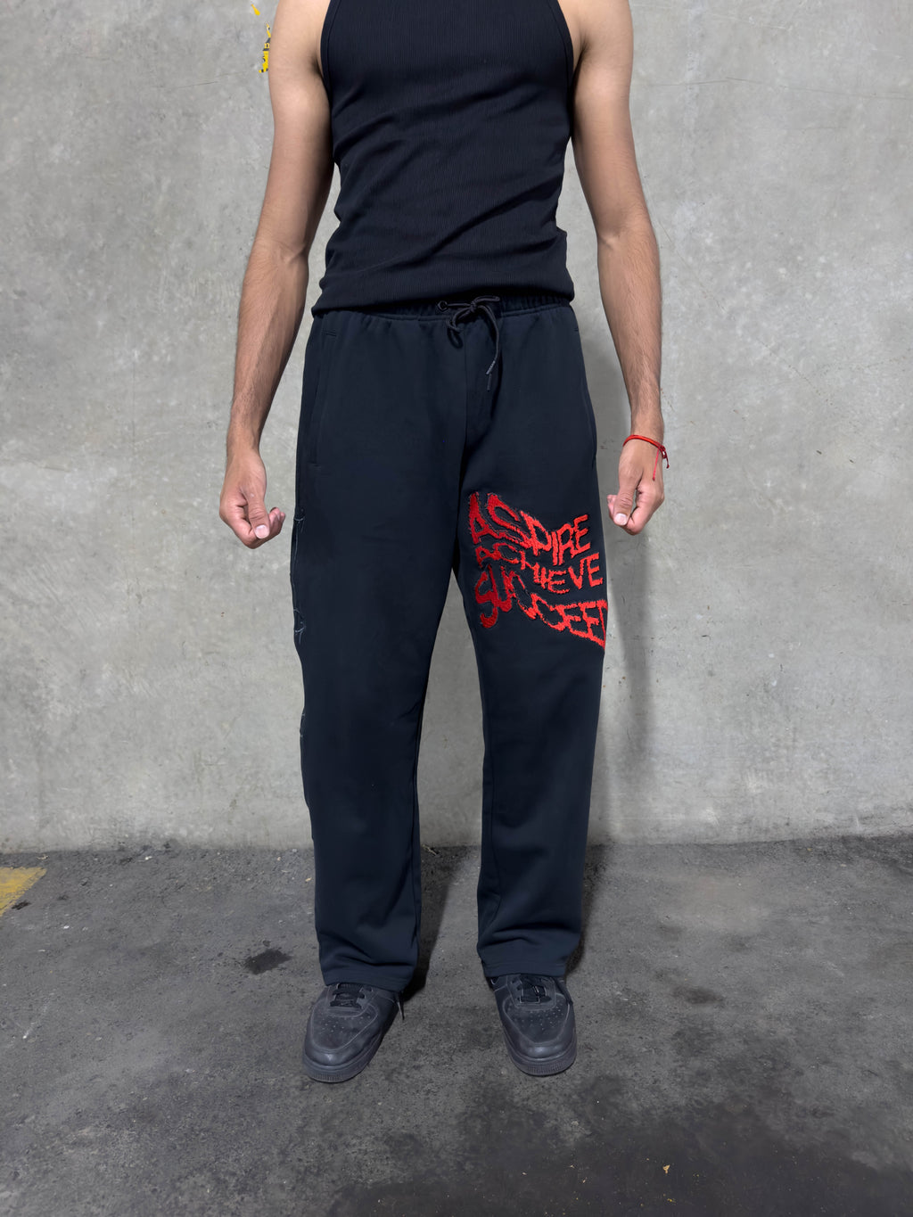 A person wearing black sweatpants with a red distressed graphic on the left thigh, paired with a black tank top and black sneakers, standing against a gray concrete wall.
