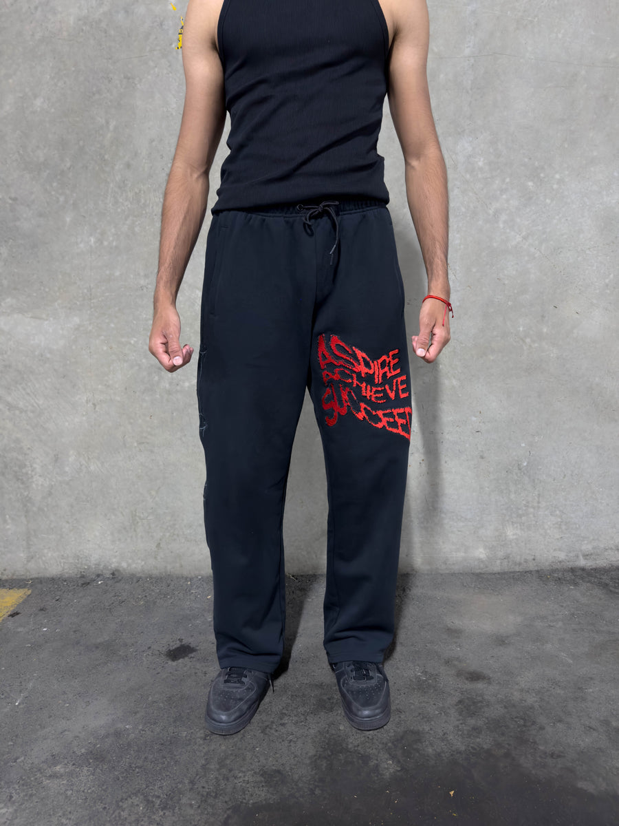 A person wearing black sweatpants with a red distressed graphic on the left thigh, paired with a black tank top and black sneakers, standing against a gray concrete wall.