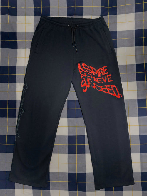 Black sweatpants with a red graphic text print on the left thigh, laid flat on a blue and white plaid blanket.