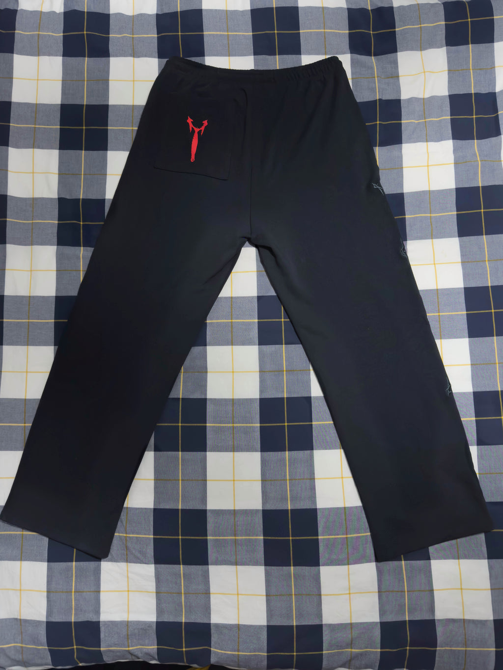 Black athletic pants with a small red kangaroo pocket and logo on the left hip, laid flat on a blue, white, and yellow plaid blanket.