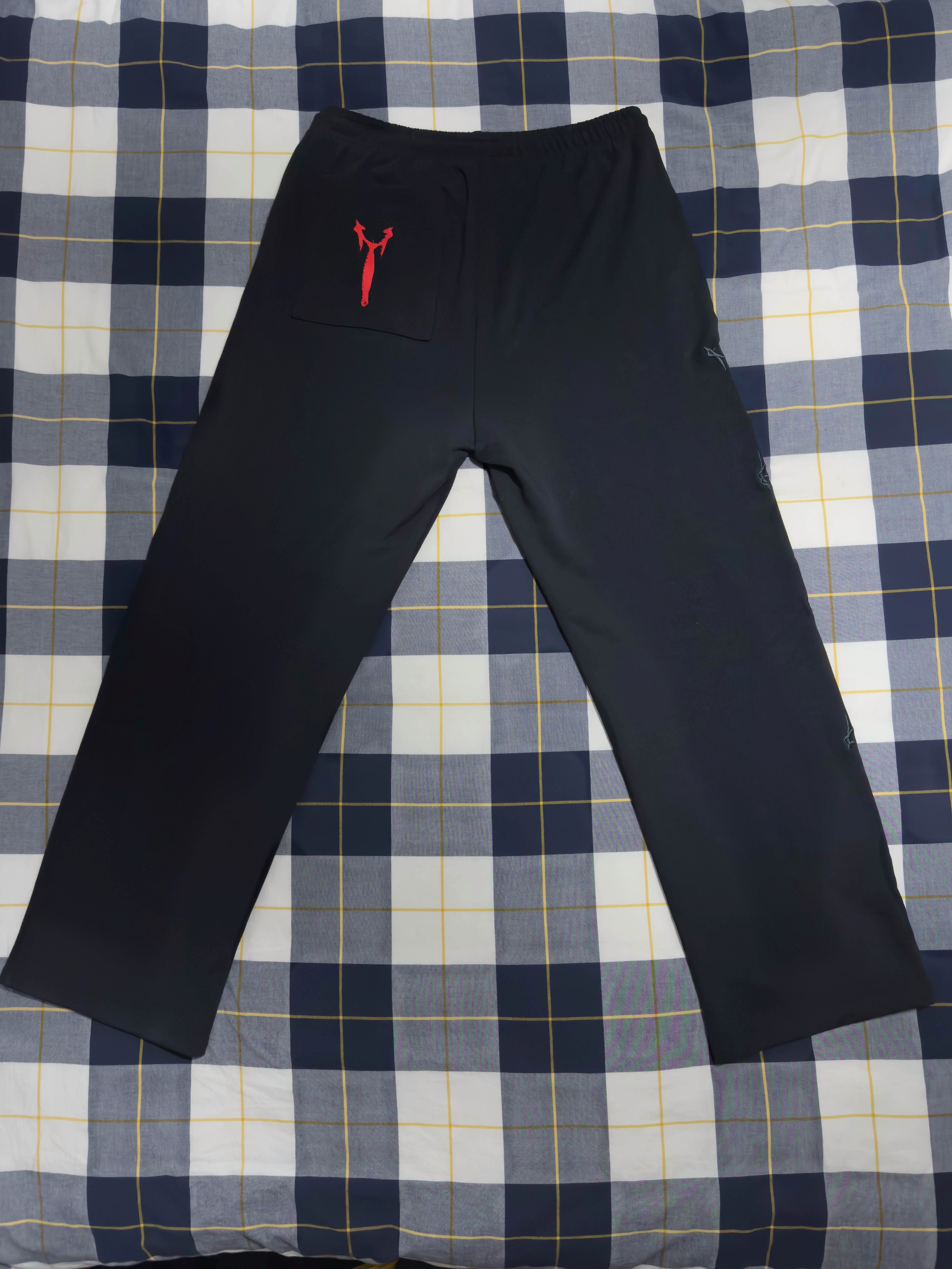 Black athletic pants with a small red kangaroo pocket and logo on the left hip, laid flat on a blue, white, and yellow plaid blanket.
