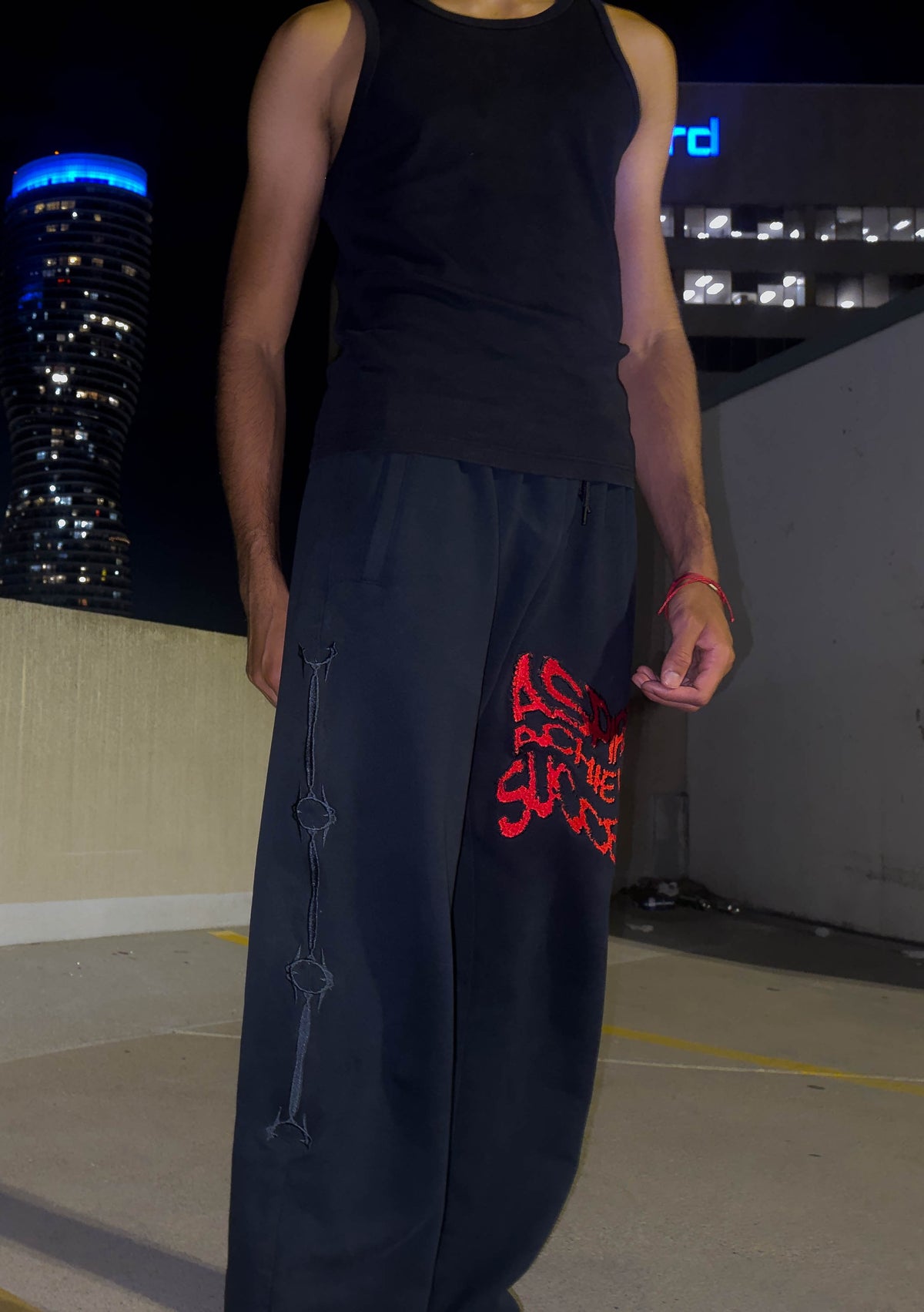 A person wearing a black tank top and very loose black pants with red embroidered lettering on one leg, standing outdoors at night with city buildings in the background.